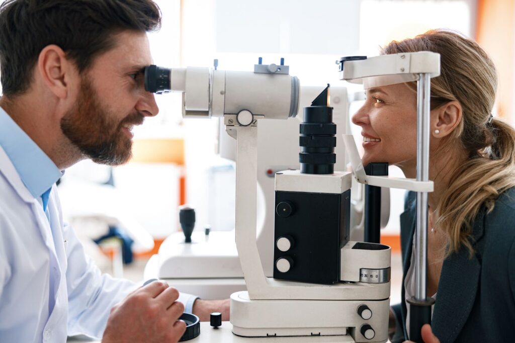 A male optometrist looks through a machine at a young woman’s eyes during her biannual comprehensive eye exam.