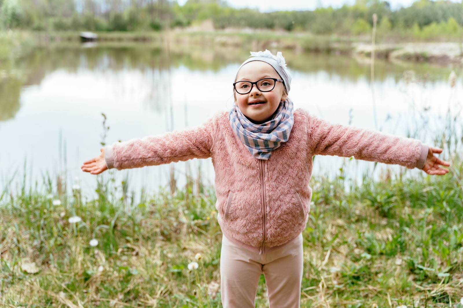 A young girl with glasses, dressed in pink, smiles and stretches her arms out while on a walk near a lake.