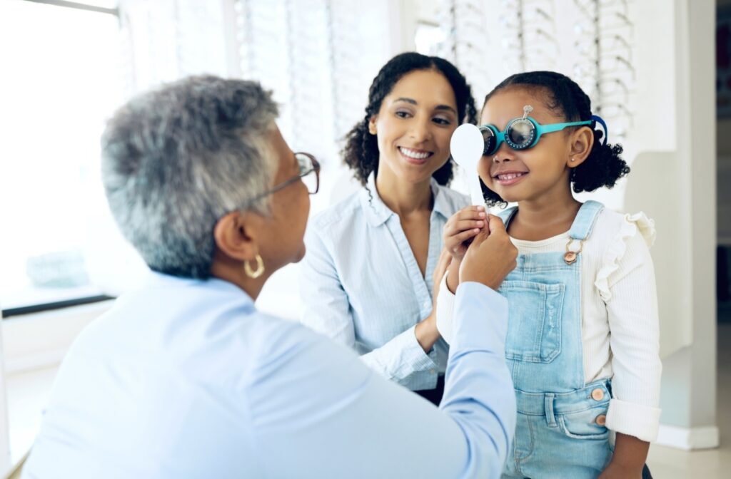 A young girl stands next to her mother in an optometry office. She smiles as the optometrist carries out an eye exam, examining her eyes with a special tool.