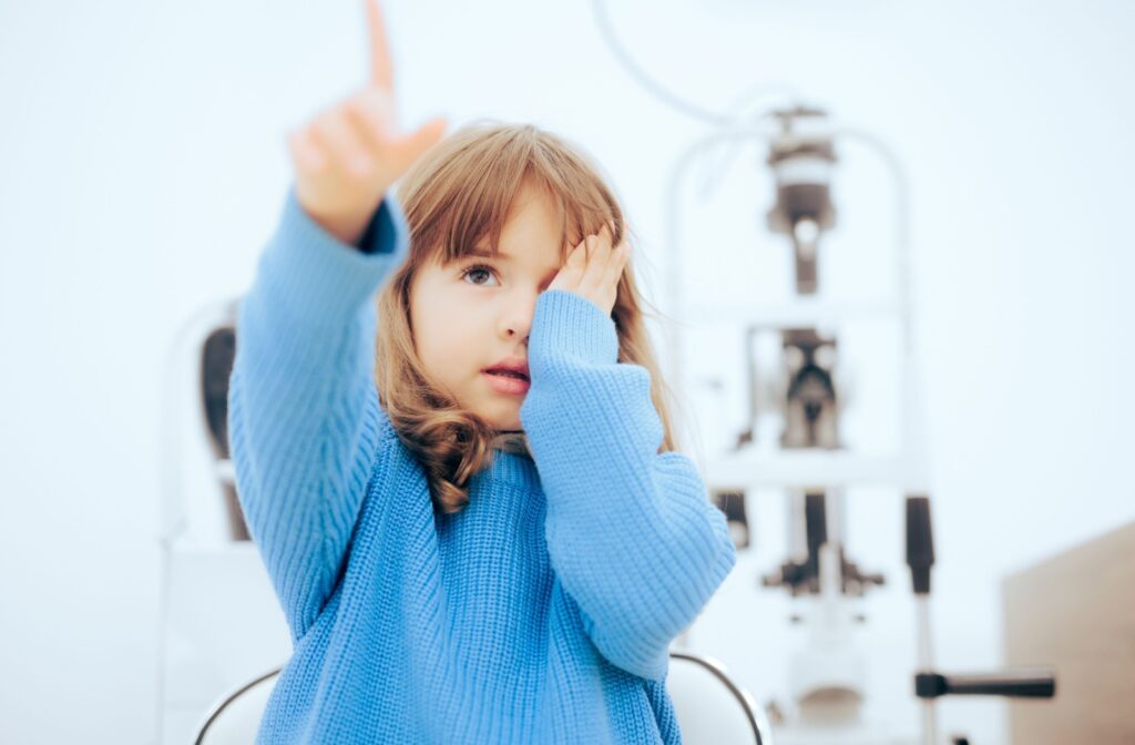 A young child in a blue knit sweater covers one eye while reading an eye exam chart to be tested for myopia.