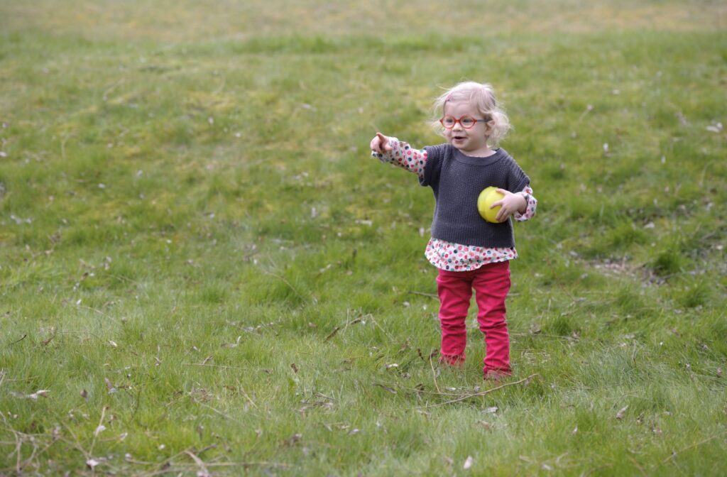 A young child wearing red glasses holds a yellow ball while playing outside to reduce the risk of myopia progression.