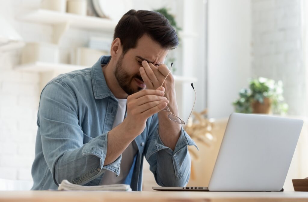 A young man sits in front of a laptop, appearing to be experiencing a headache.