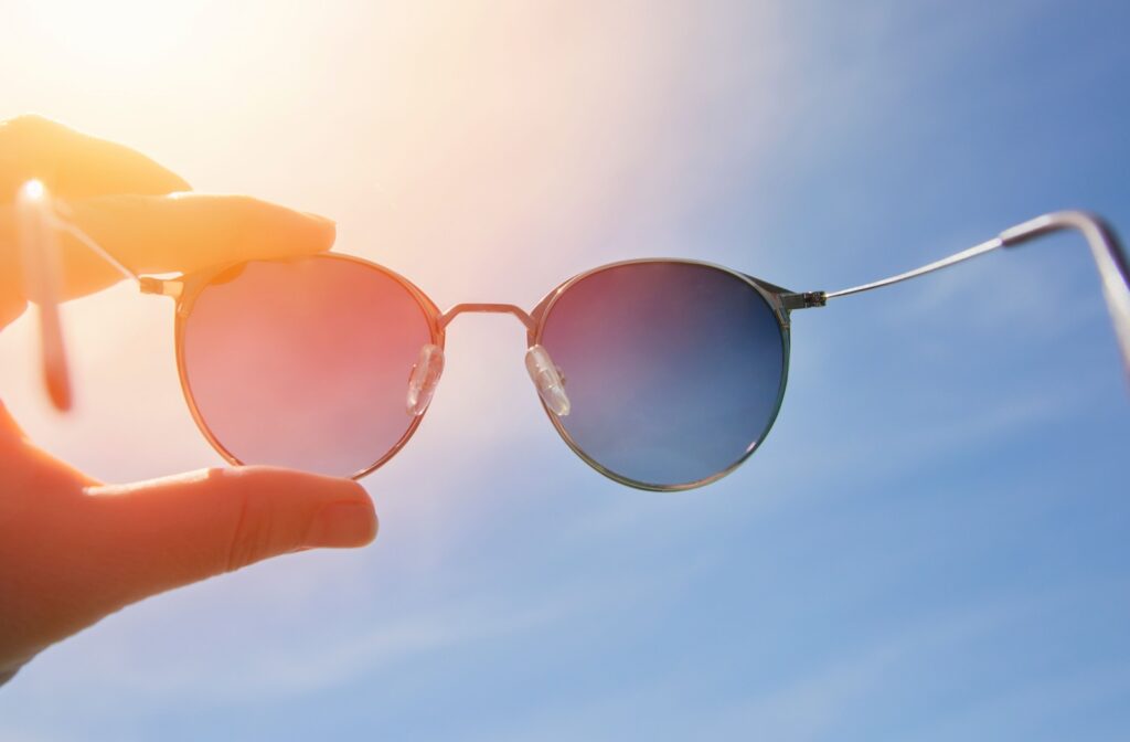 A person holds up a pair of sunglasses toward a bright sun with a clear blue sky in the background.