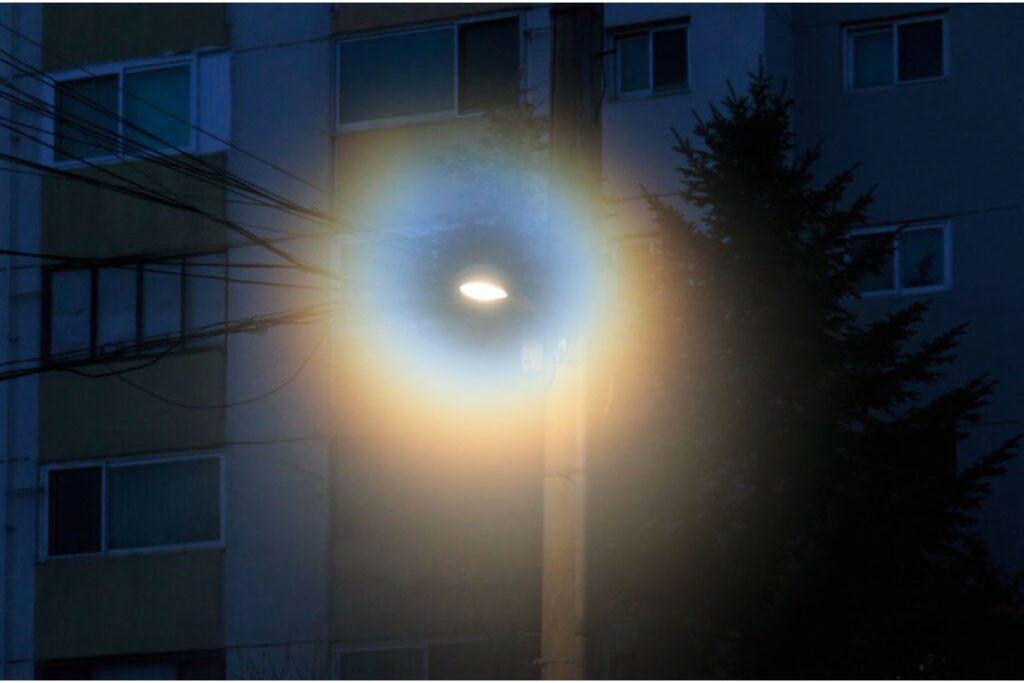 A photograph taken at night showing a street lamp glowing against a dark apartment building. The light from the lamp creates a large, blurry, hazy halo effect around the light source, illustrating the visual symptom of halos, which can indicate conditions like cataracts or corneal issues.